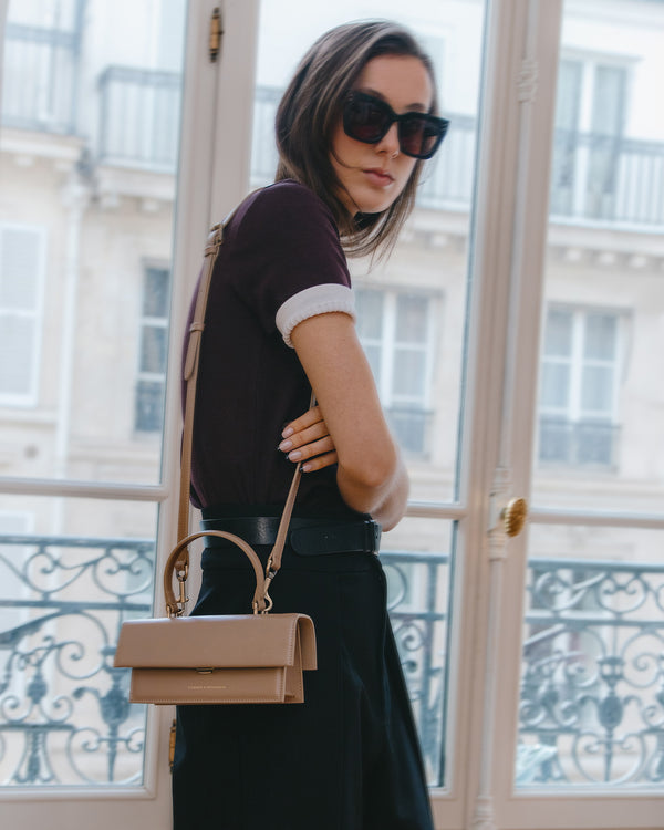 THEA MINI bag in beige worn crossbody, side view framed by a Parisian window
