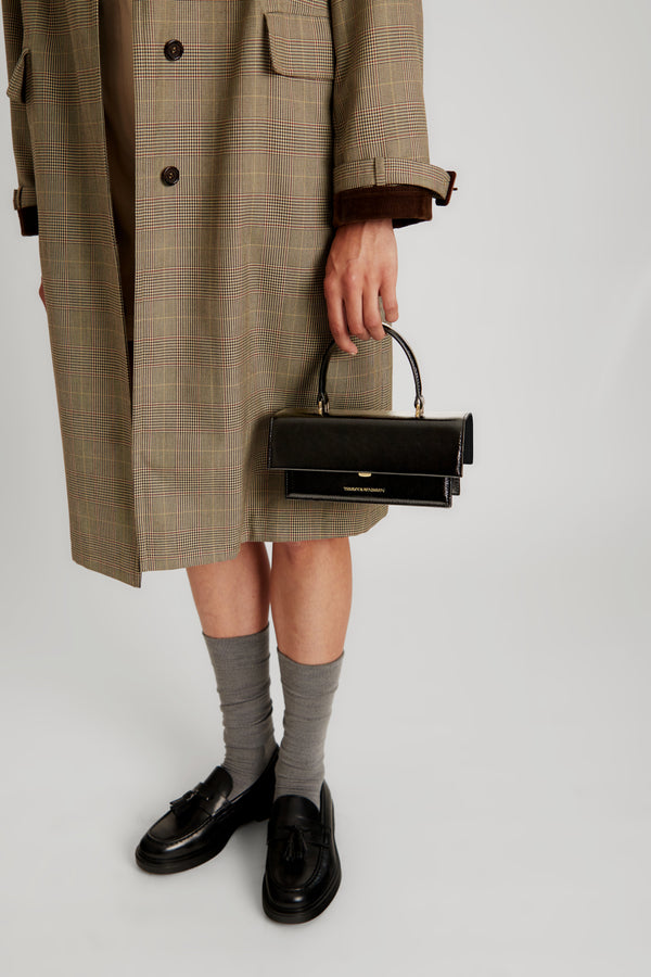 THEA MINI bag in black naplak carried by hand, clasp detail, held by a person in a Prince of Wales check coat