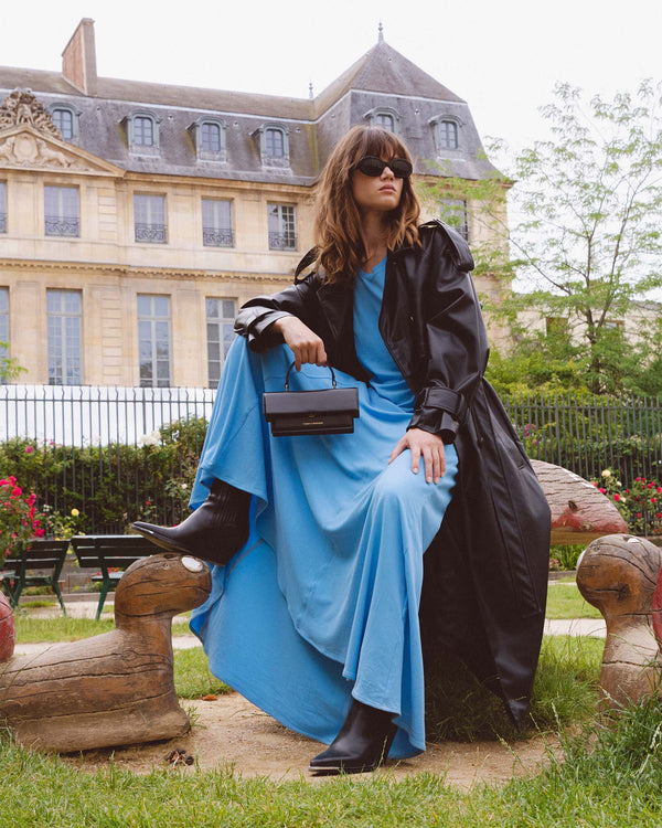 THEA MINI bag in black held by a woman seated in a blue dress and black trench coat