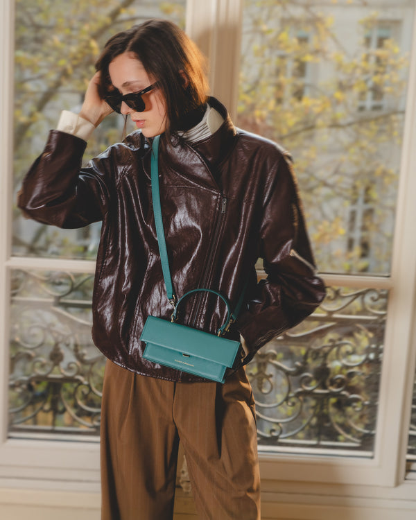 THEA MINI bag in Turquoise worn crossbody, brown leather jacket, Parisian window background